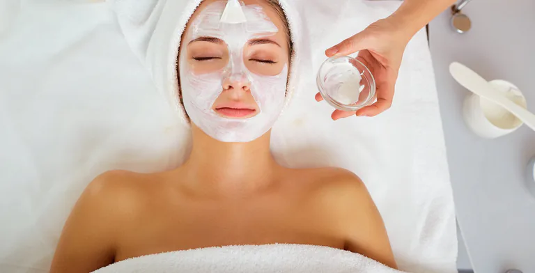A client in a towel turban rests, eyes closed, under a cream mask, a glass bowl of cream beside her.