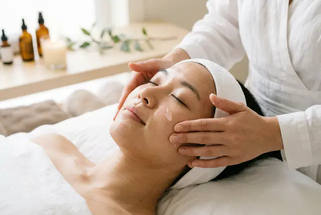 Hands cup a relaxed client's cheeks to press in serum, amber bottles and a candle on the sill behind.