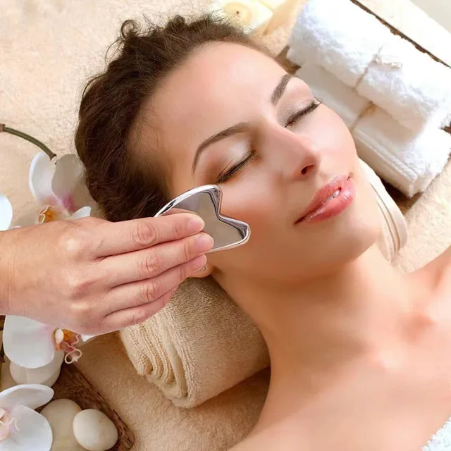 A heart-shaped steel gua sha tool glides along the cheekbone of a smiling client, white orchids and pebbles beside her.
